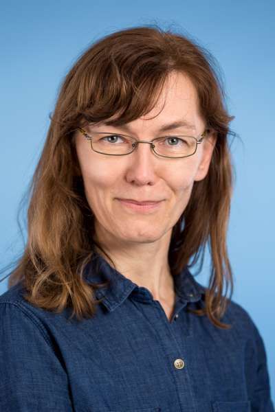 Eine Person mit schulterlangem braunem Haar und Brille, die ein dunkelblaues Hemd vor einem einfarbig hellblauen Hintergrund trägt.
