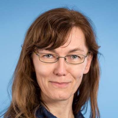 Ein Porträt einer Person mit schulterlangem braunem Haar und Brille, die vor einem schlichten blauen Hintergrund leicht lächelt.