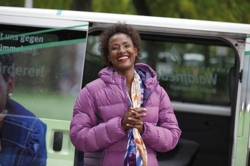 Eine lächelnde Person, die eine lila Daunenjacke und einen bunten Schal trägt, steht vor einem weißen Fahrzeug.