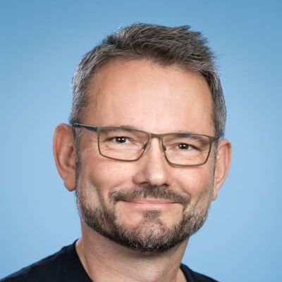 Eine Person mit kurzen, dunklen Haaren und Bart, die eine Brille trägt, lächelt vor einem einfarbig blauen Hintergrund.