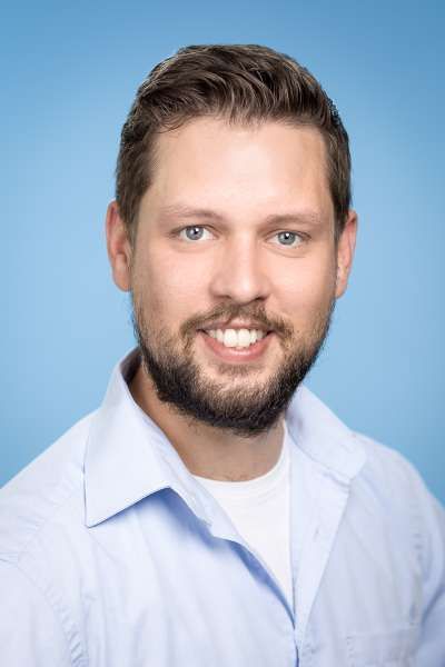 Ein Mann mit Bart trägt ein blaues Hemd und lächelt in die Kamera.
