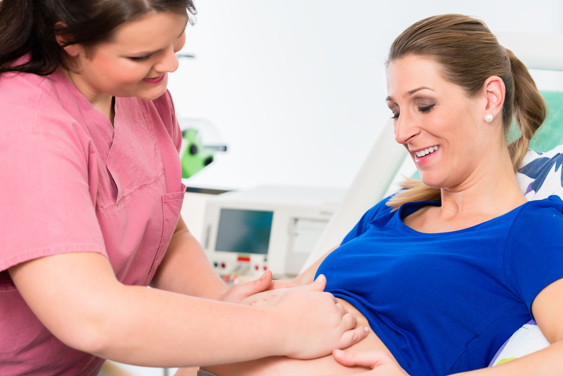 Eine Krankenschwester untersucht den Bauch einer schwangeren Frau in einer medizinischen Einrichtung; beide lächeln.