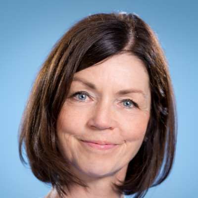 Ein Porträt einer Person mit schulterlangem braunem Haar und blauen Augen, die vor einem schlichten blauen Hintergrund leicht lächelt.