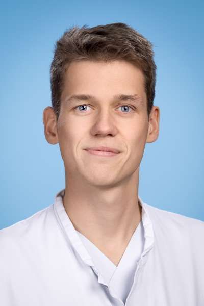 Ein junger Mann im weißen Laborkittel lächelt in die Kamera.