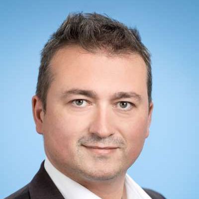 Porträt eines Mannes mit kurzen braunen Haaren, der einen dunklen Anzug und ein weißes Hemd trägt, vor hellblauem Hintergrund.