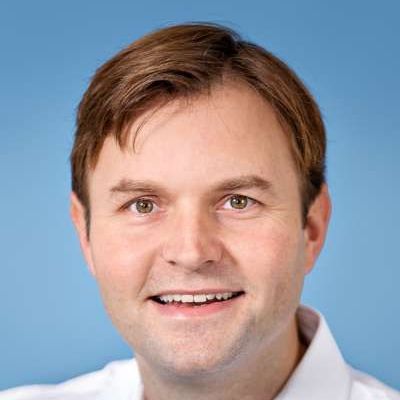 Porträt einer Person mit braunen Haaren, die vor einem einfarbig blauen Hintergrund lächelt.