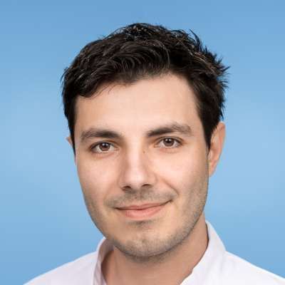 Ein Mann in einem weißen Hemd lächelt vor einem blauen Hintergrund in die Kamera.