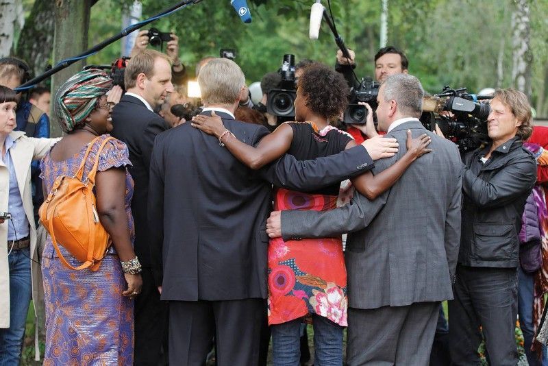 Eine Gruppe von Menschen in formeller Kleidung steht Arm in Arm beieinander, umgeben von Kameras und Mikrofonen.
