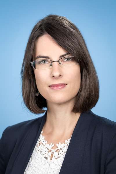 Eine Frau mit Brille und Jacke steht vor einem blauen Hintergrund.