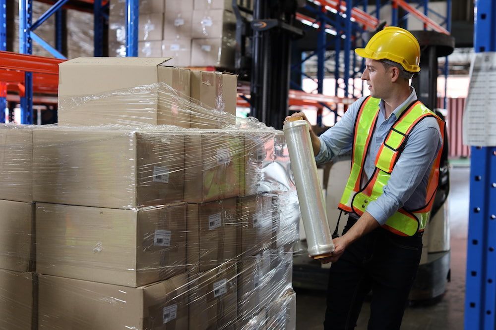 Warehouse worker wrapping boxes with plastic wrap, wearing hard hat and safety vest.