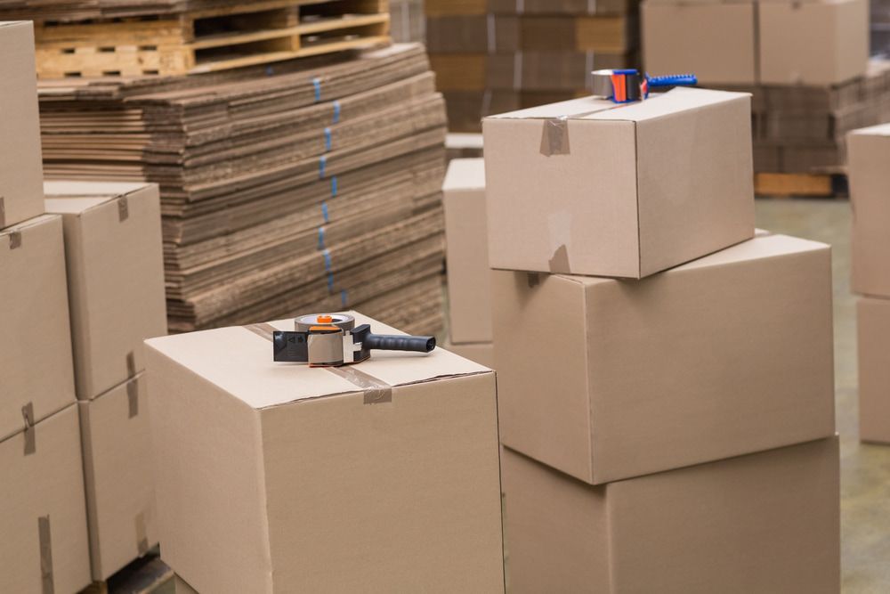 Cardboard boxes stacked in a warehouse, some with tape dispensers on top, and cardboard sheets in the background.