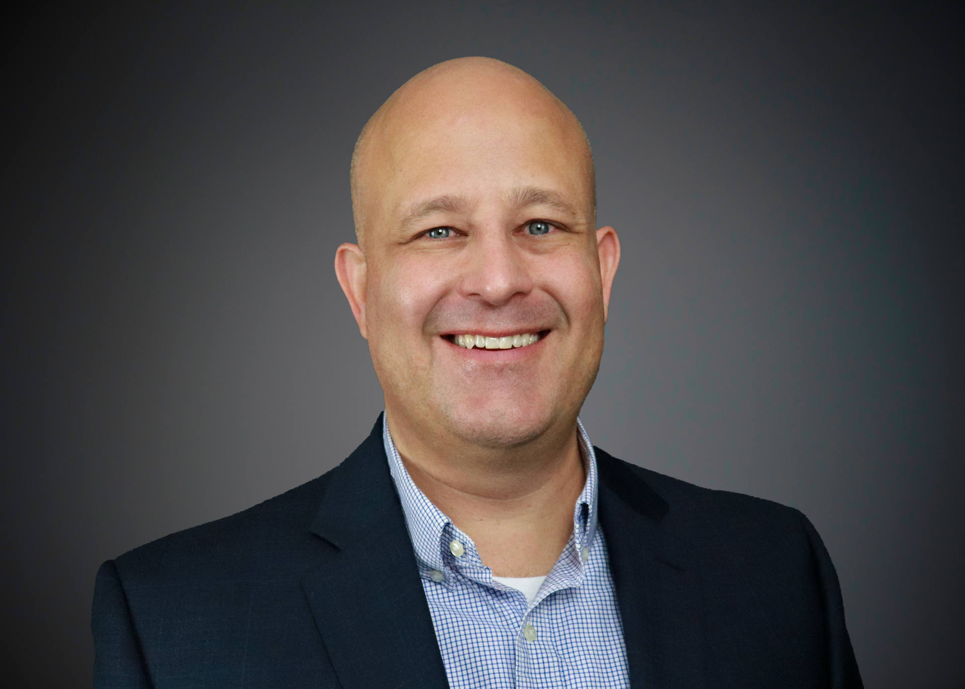 Smiling bald man in a dark blazer and patterned shirt against a gray studio background