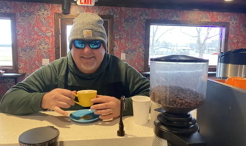 Smiling person in a knit hat and sunglasses holds a yellow cup at a cafe table beside a coffee grinder.
