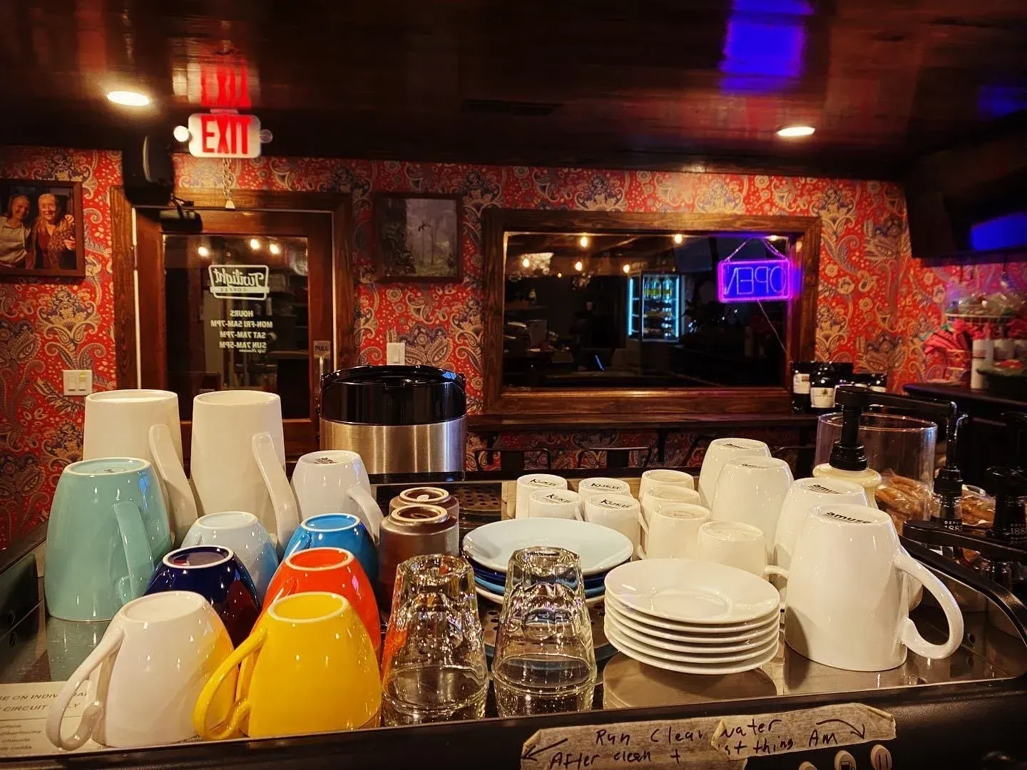 Rows of colorful mugs and stacked dishes on a bar counter in a dimly lit restaurant or cafe.