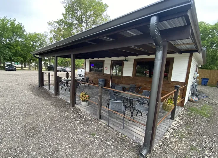 Covered outdoor patio with black metal posts, tables, chairs, and a gravel yard beside a building