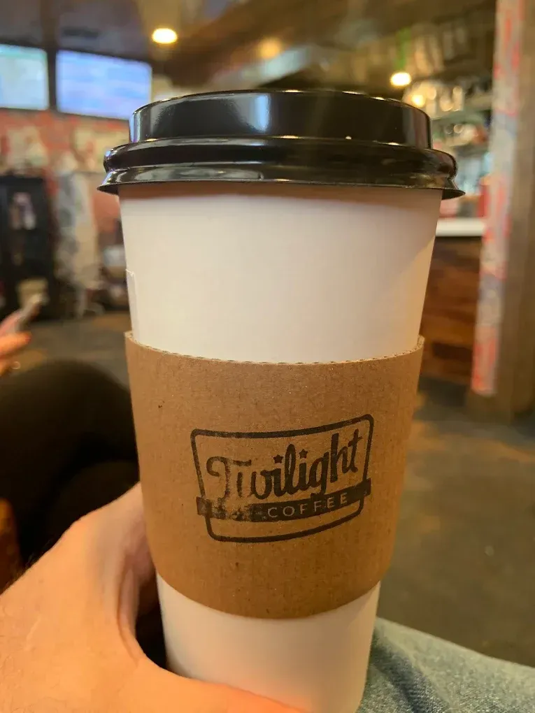 White takeaway coffee cup with black lid and brown sleeve in a hand, in a café setting