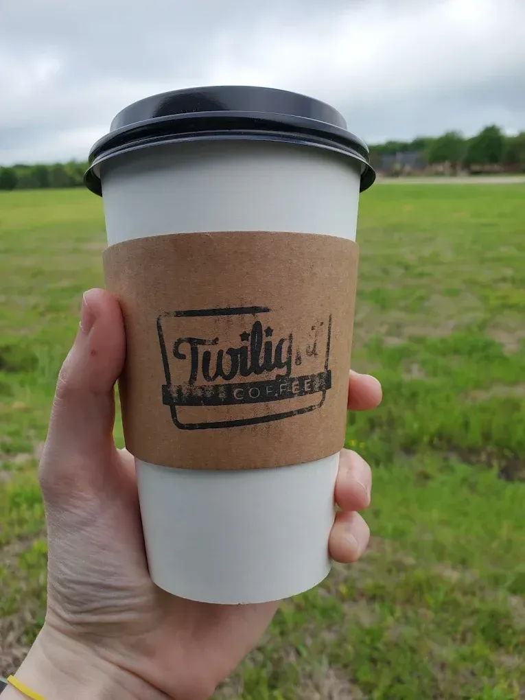 Hand holding a white coffee cup with a brown sleeve and black lid outdoors in a grassy field