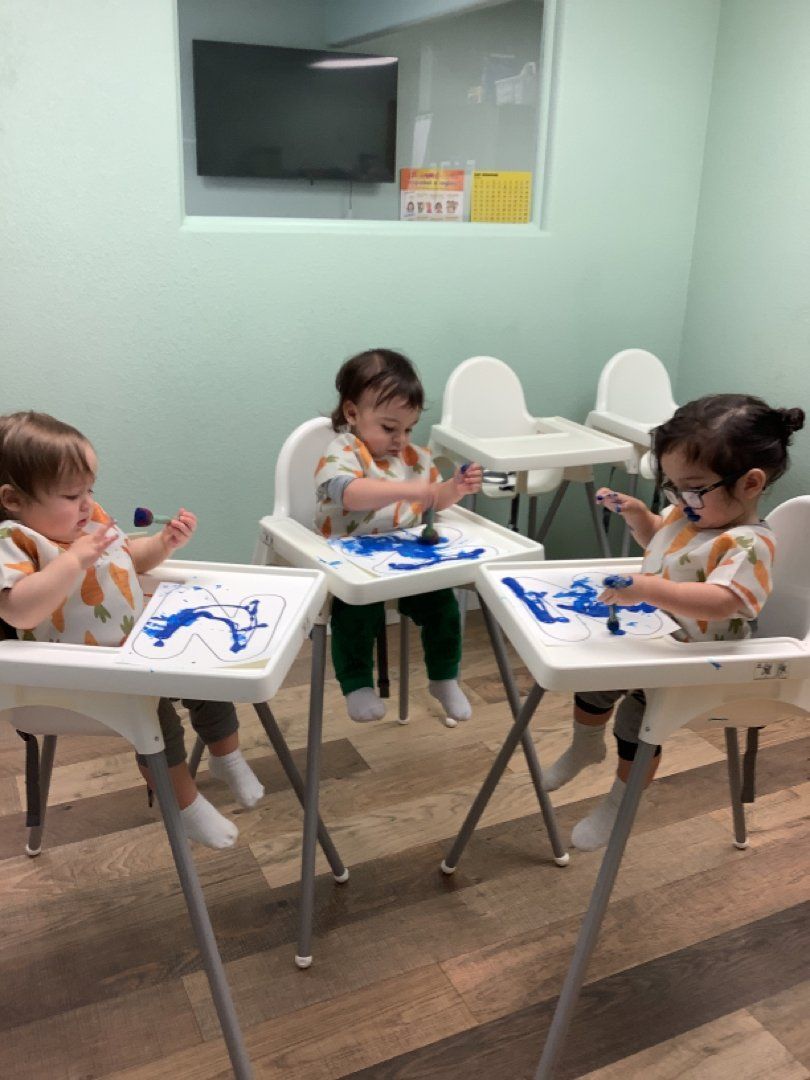 Three toddlers are sitting in high chairs playing with blue paint.