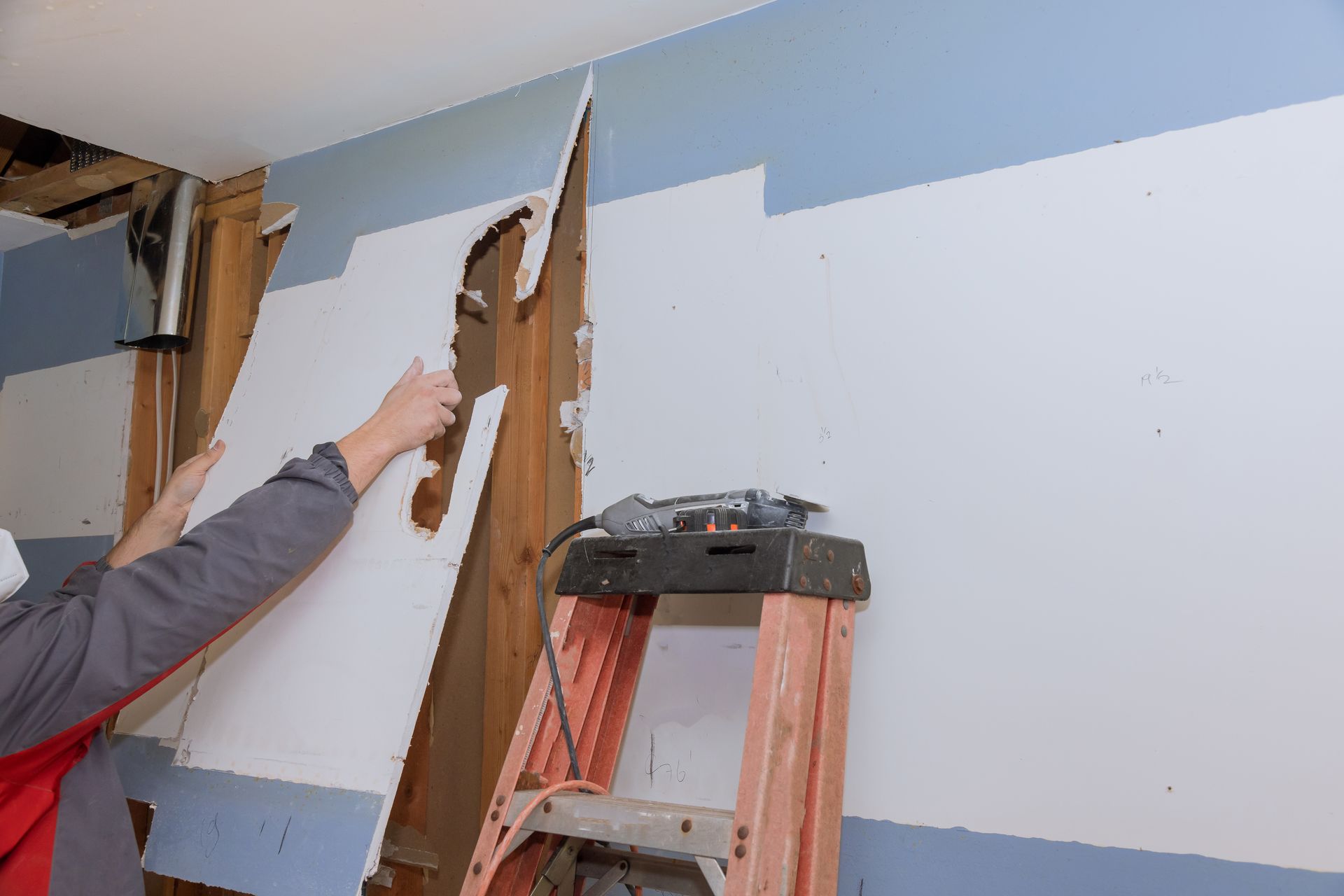 Person removing drywall panels inside a room with a ladder and exposed wall studs