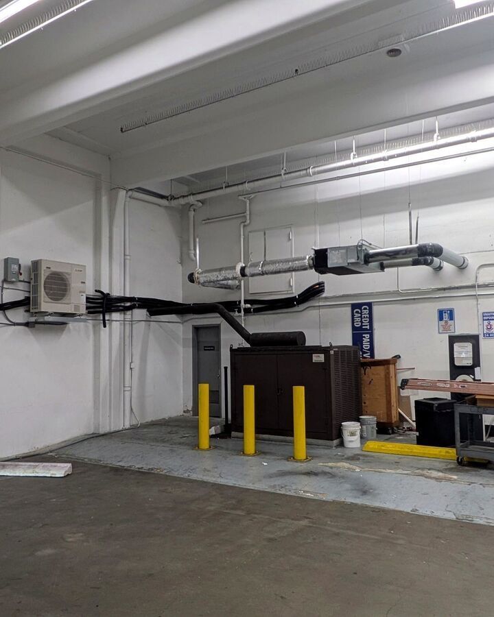 Industrial storage area with yellow bollards, ductwork, and a dark equipment cabinet against a white wall.