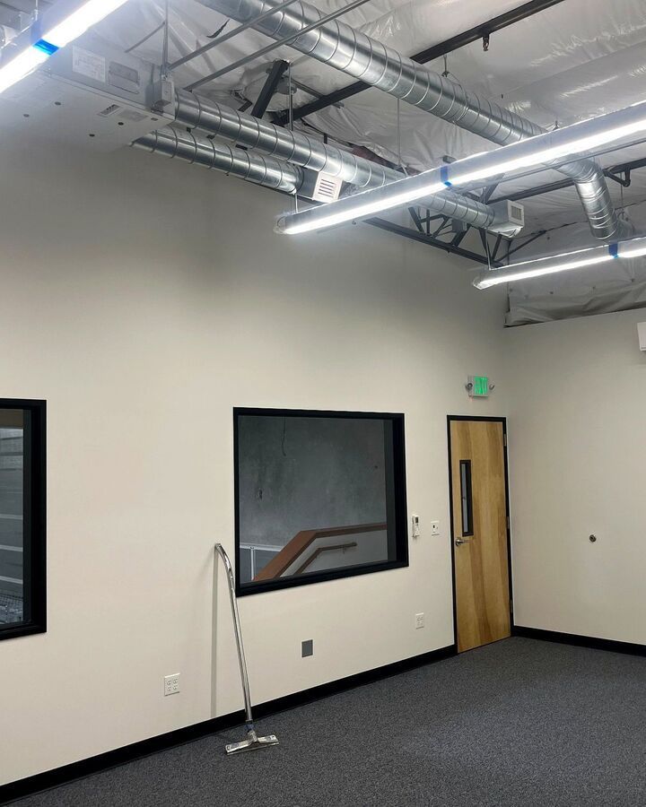 Empty office room with white walls, black carpet, large interior windows, and exposed ceiling ducts and lights