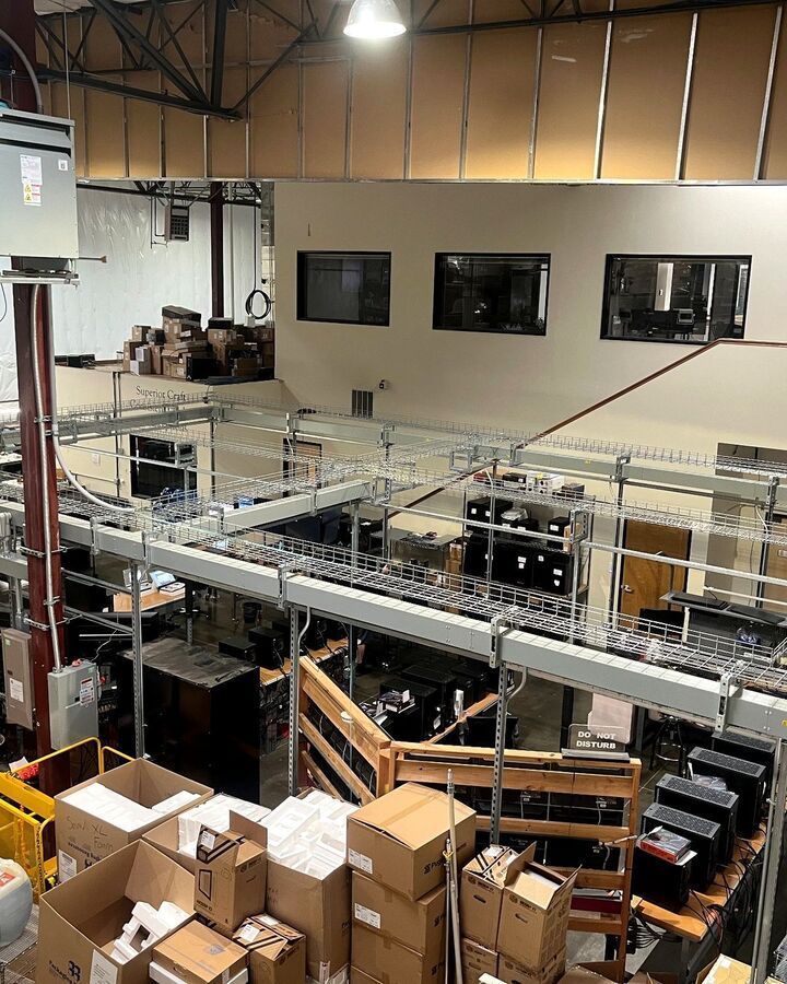 Warehouse interior with conveyor belts, stacked cardboard boxes, and mezzanine office windows.