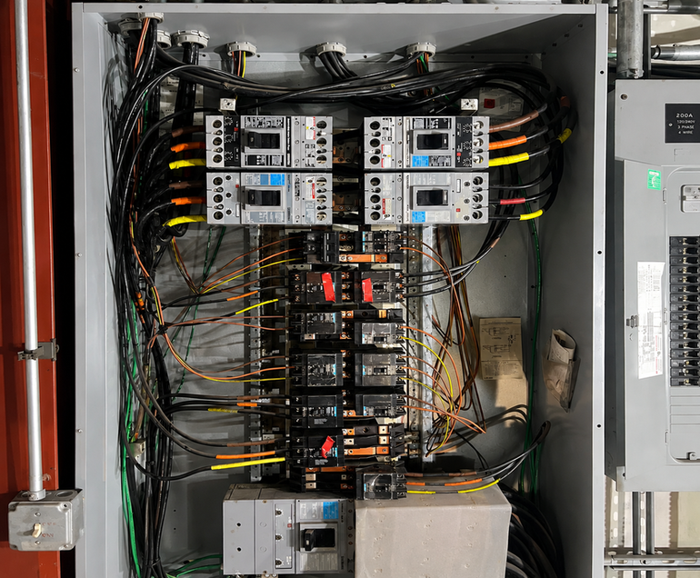 Open electrical control panel with breakers, relays, and tangled wiring inside a metal cabinet.