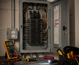 Open electrical breaker panel with wiring, outlet, and tools on a workbench in a utility room
