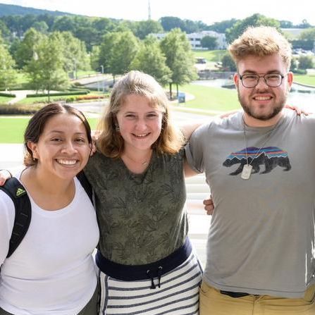 Three smiling friends standing outdoors on a sunny day with a green park in the background