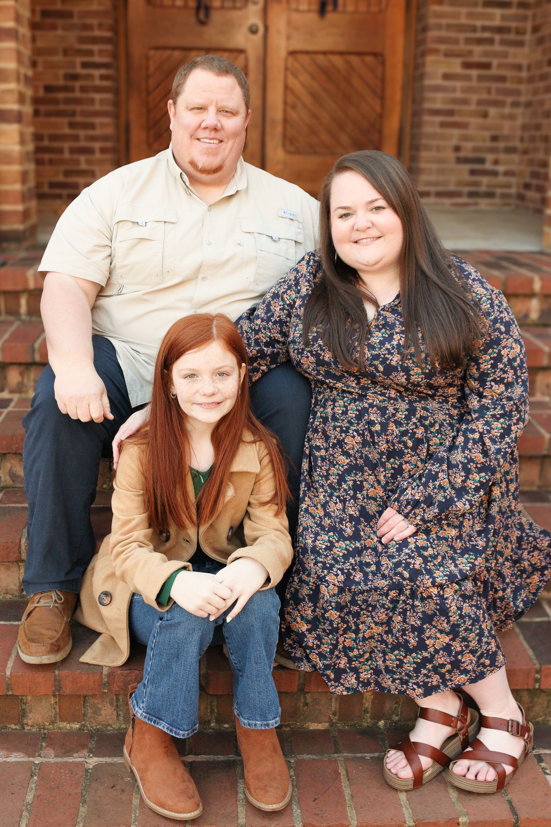 Three people sitting on brick steps in front of a brick house, smiling at the camera.