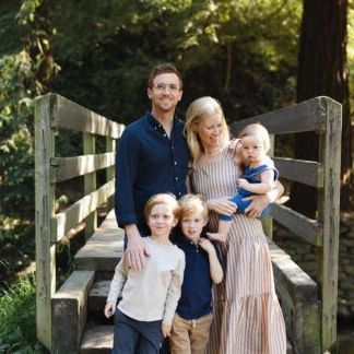 Family of five standing on a wooden bridge in a wooded park, smiling at the camera.
