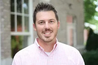 Smiling man in a light pink striped shirt standing outdoors by a building