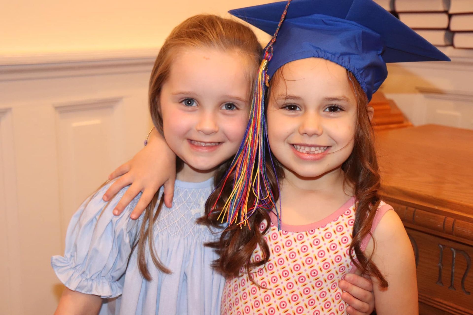 Two smiling children posing indoors, one wearing a blue graduation cap and holding the other close