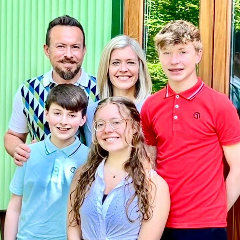Five people smiling outdoors in front of a green wooden wall, wearing colorful shirts.