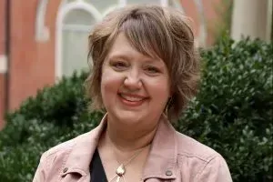 Smiling woman in a pink jacket standing outdoors with greenery and a brick building behind her