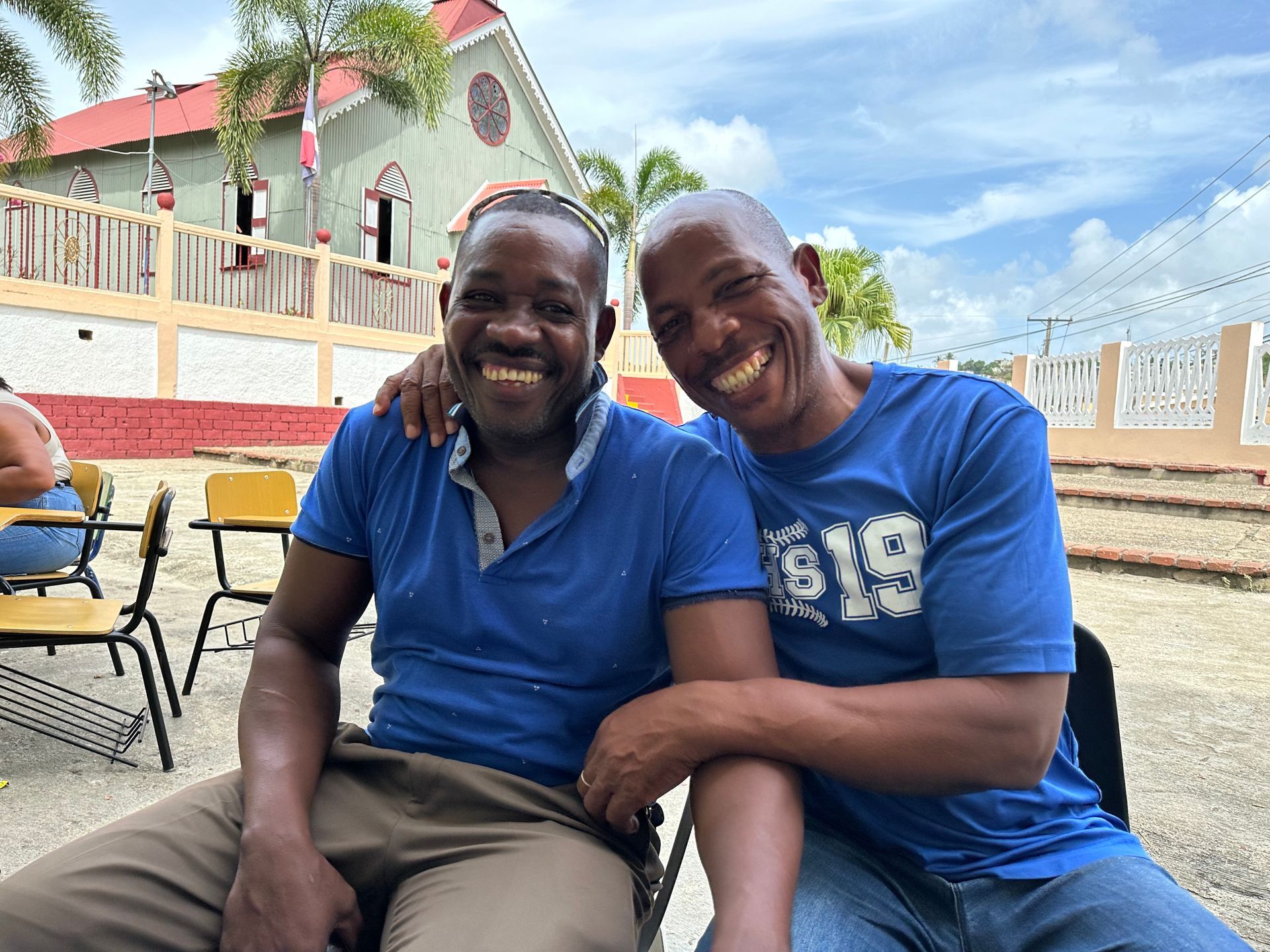 Two smiling people in blue shirts sitting together outdoors on a bench near a white building.