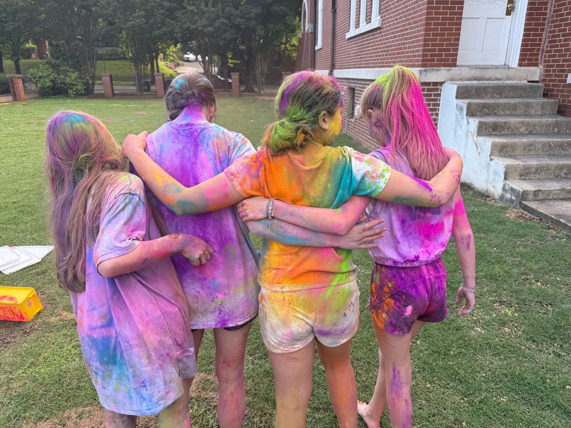 Four friends covered in colorful powder stand with arms around each other outside a brick building.