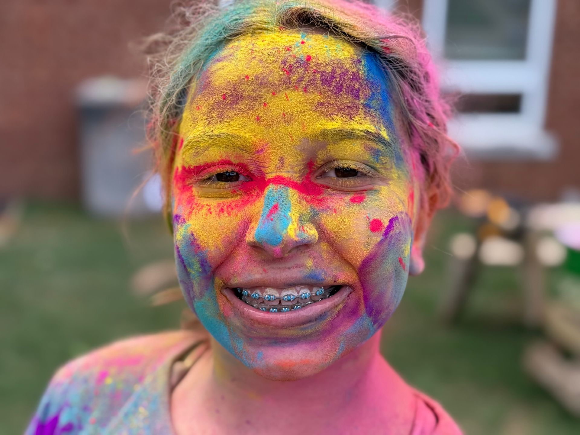 Smiling child covered in bright pink, yellow, and blue powder outdoors in a yard