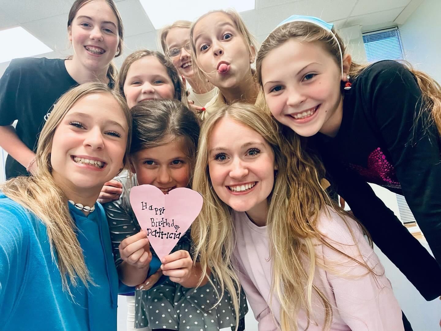 Smiling group selfie indoors, holding a pink heart sign, with one person playfully puckering lips.