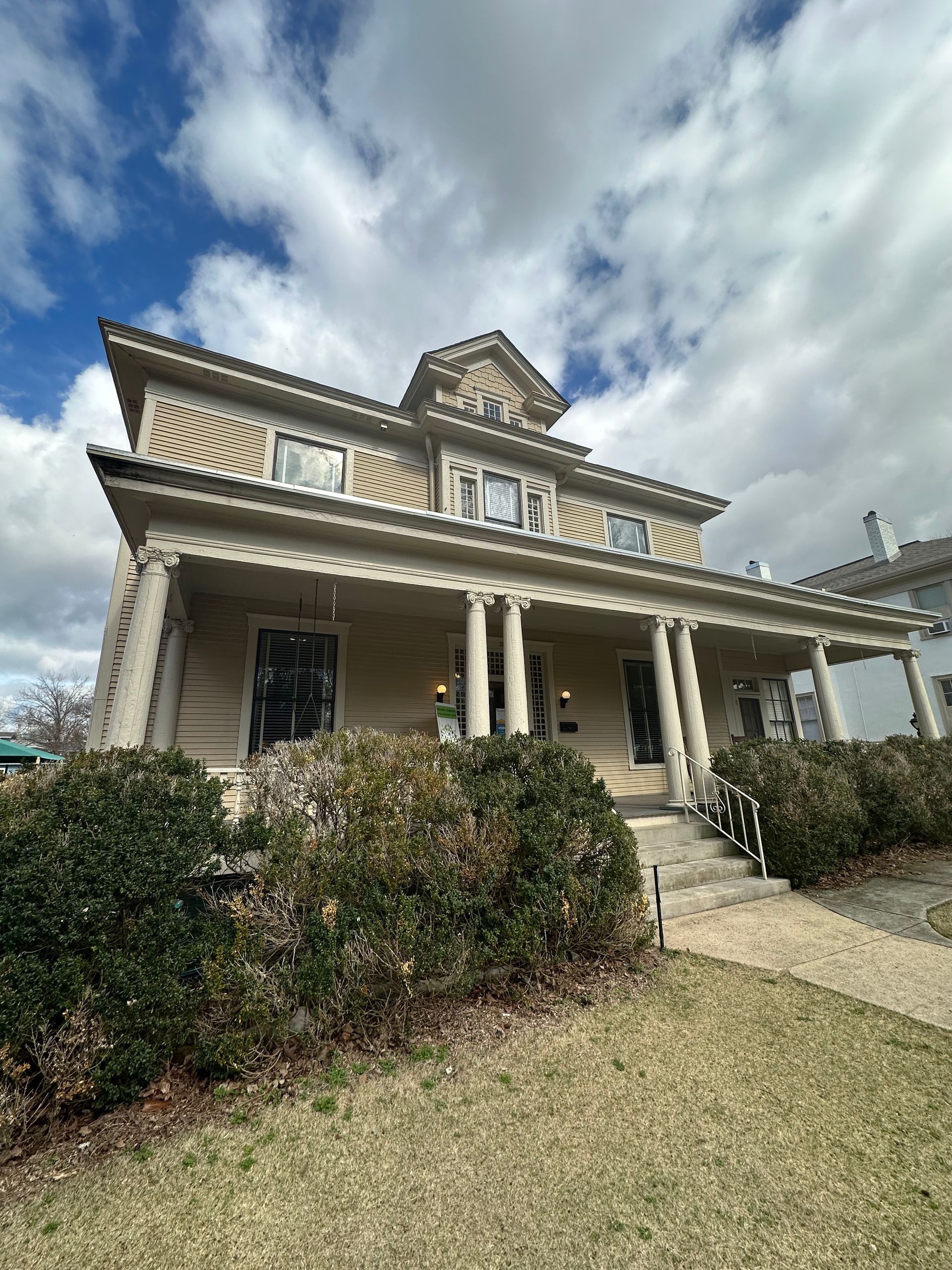 Victorian-style house with a porch, striped awning, and hedges under a cloudy sky