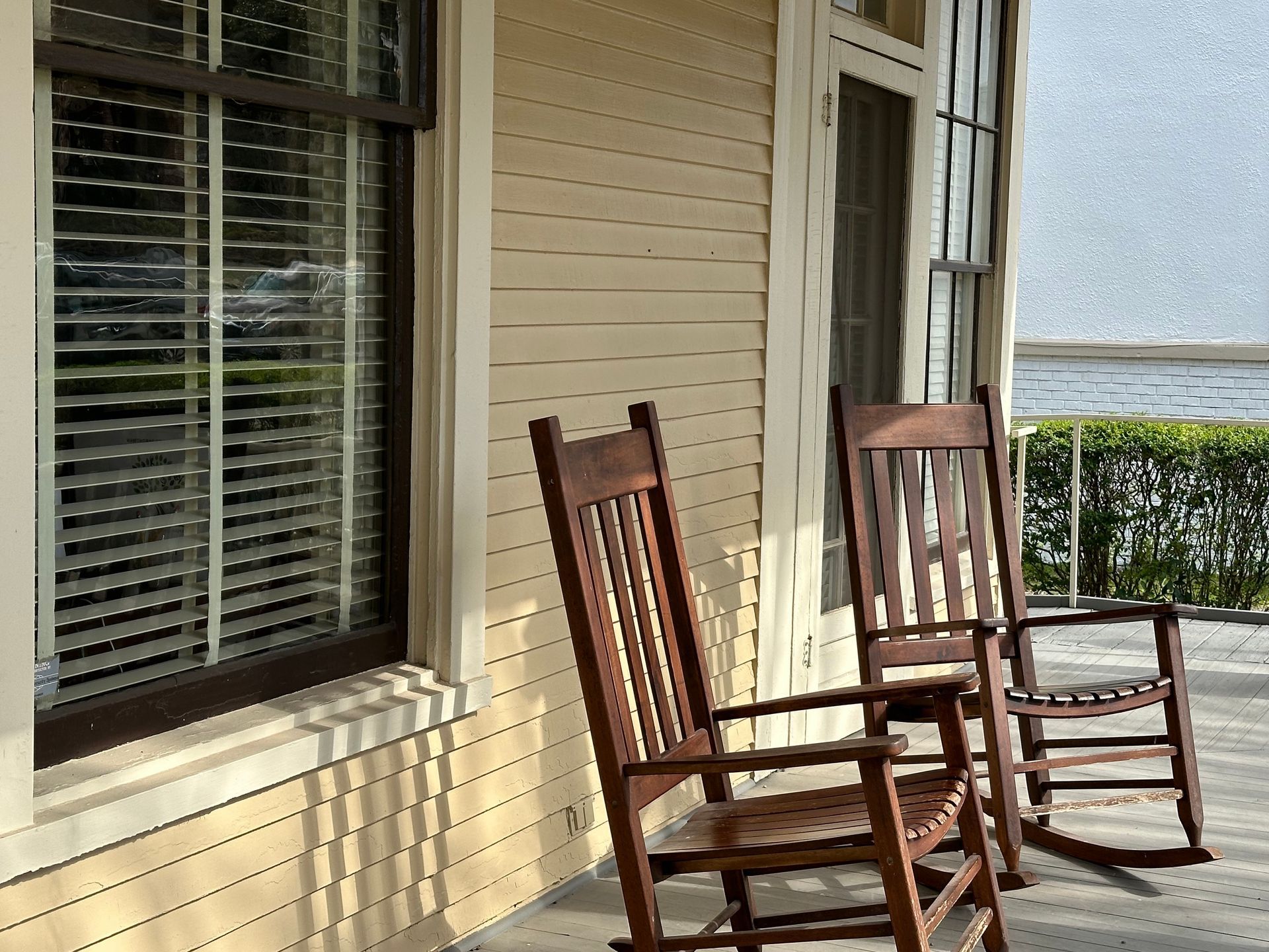 Two wooden rocking chairs on a sunlit porch beside a window and beige siding.