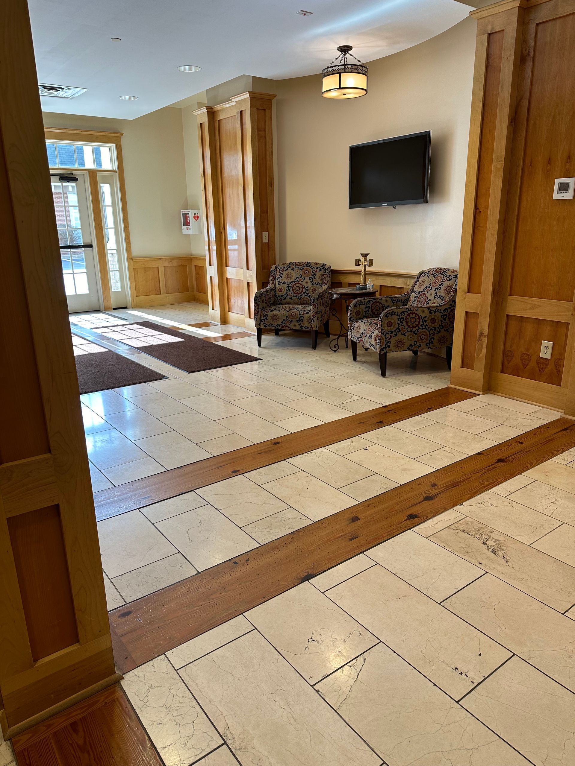 Hotel lobby with tiled floor, chairs, wall-mounted TV, and bright entrance in the background