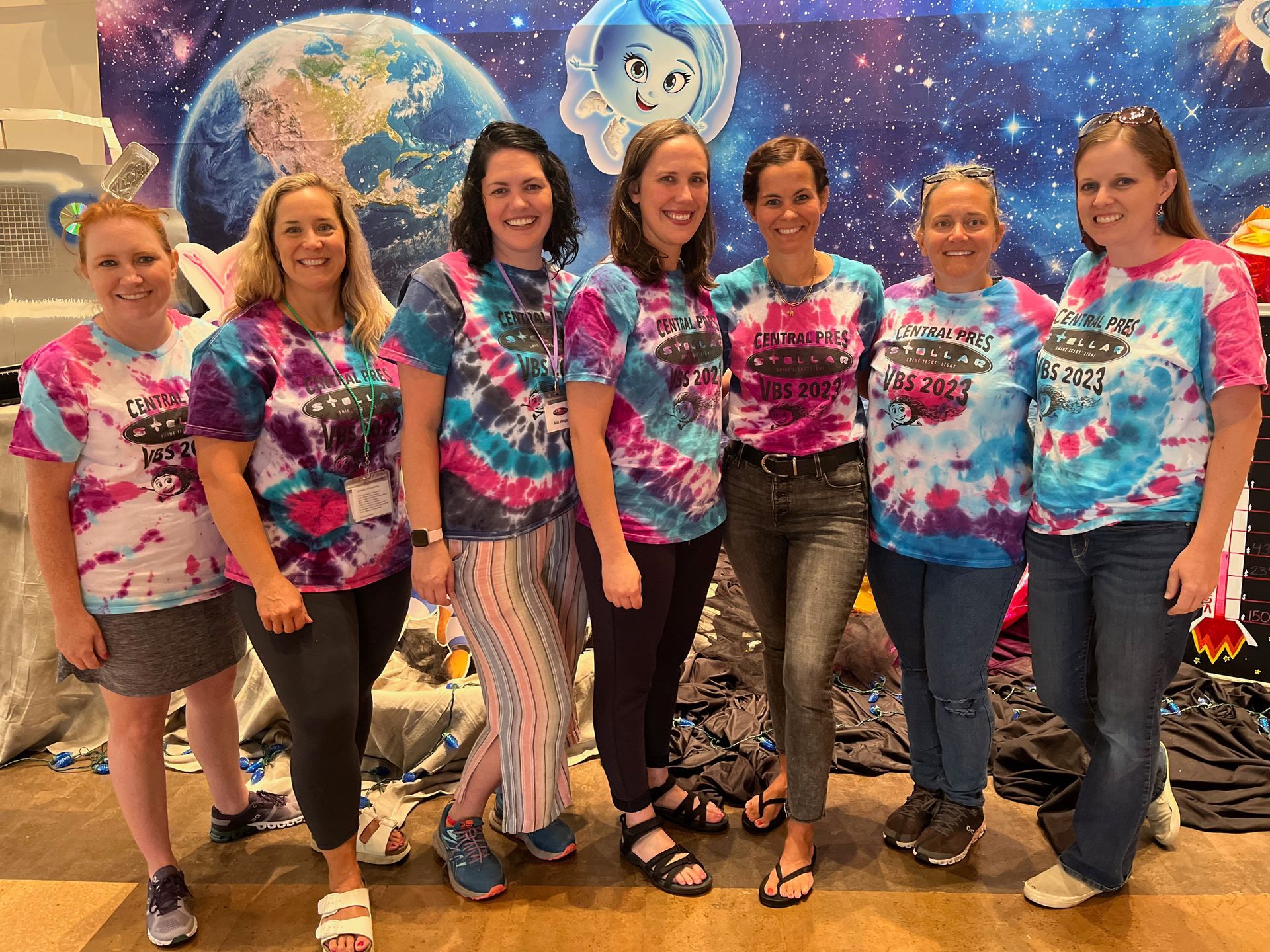 Group of seven smiling people in colorful tie-dye shirts posing in front of a starry space-themed backdrop.