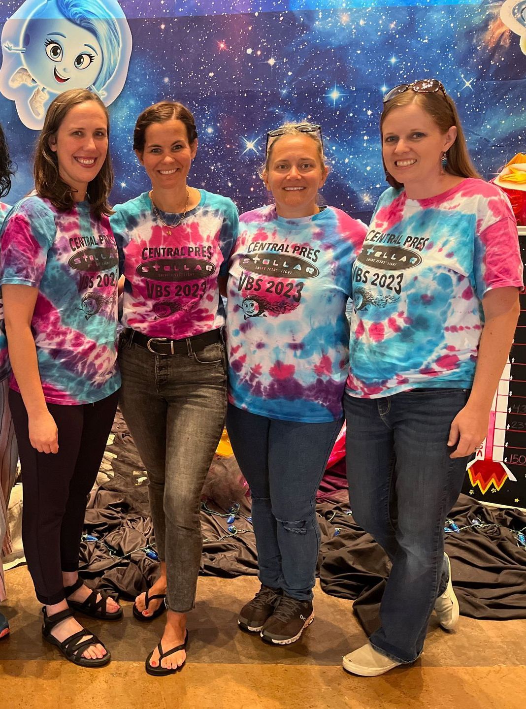 Four people in pink and blue tie-dye shirts pose together at an indoor event booth.