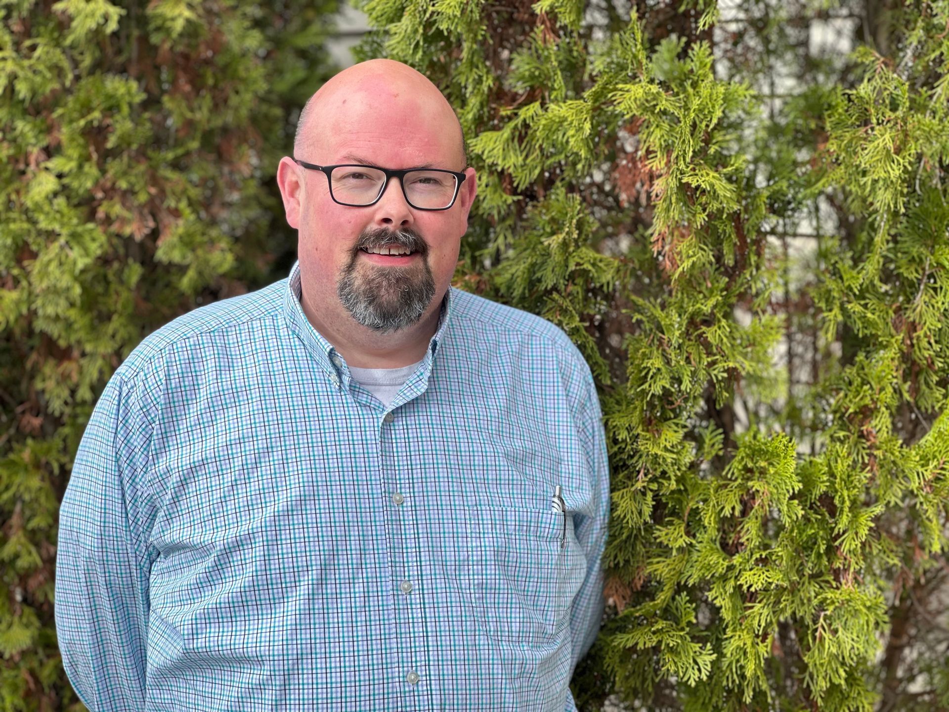 Bald man with glasses and a goatee standing before a green hedge, wearing a blue patterned shirt.