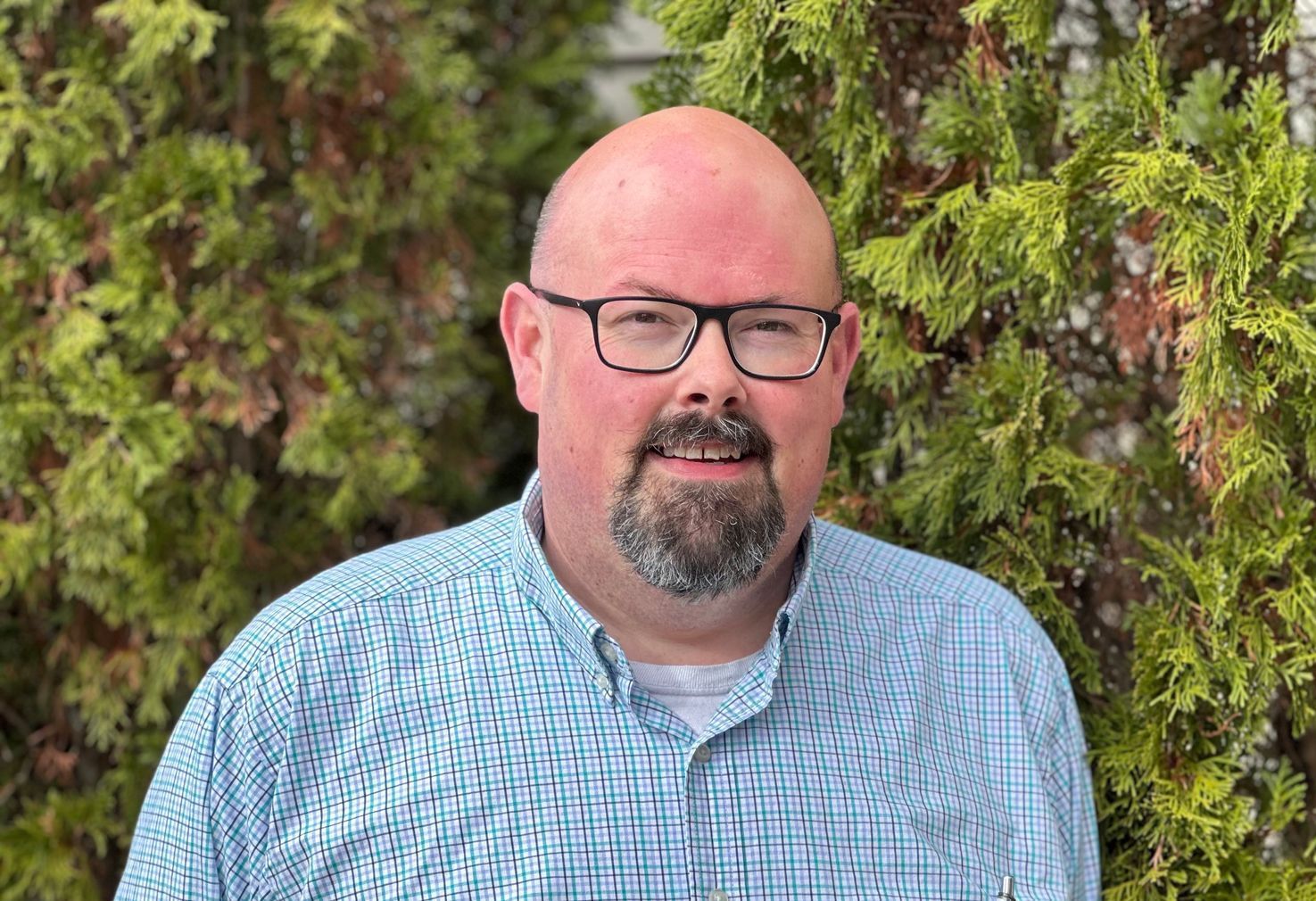 Man with glasses and goatee in a blue checkered shirt standing before green foliage