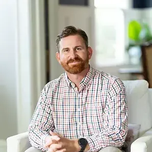 Man sitting on a white chair in a bright room, wearing a plaid shirt and looking at the camera.