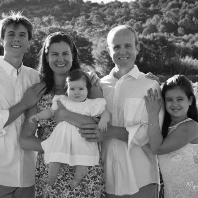 Smiling family of five posing outdoors in front of a hillside, with a baby held at center