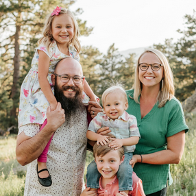 Four smiling people pose outdoors in a grassy field with trees in the background, two children on adults' shoulders.