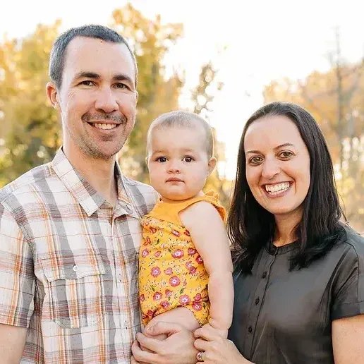 Three people smiling outdoors, with a toddler in a yellow dress held between two adults.